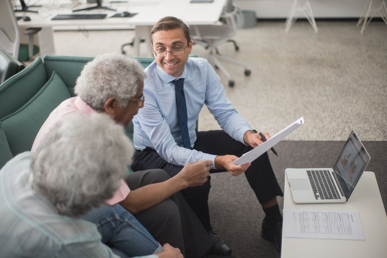 about-03 Consultant discussing financial plans with senior clients in a modern office setting, using documents and a laptop.