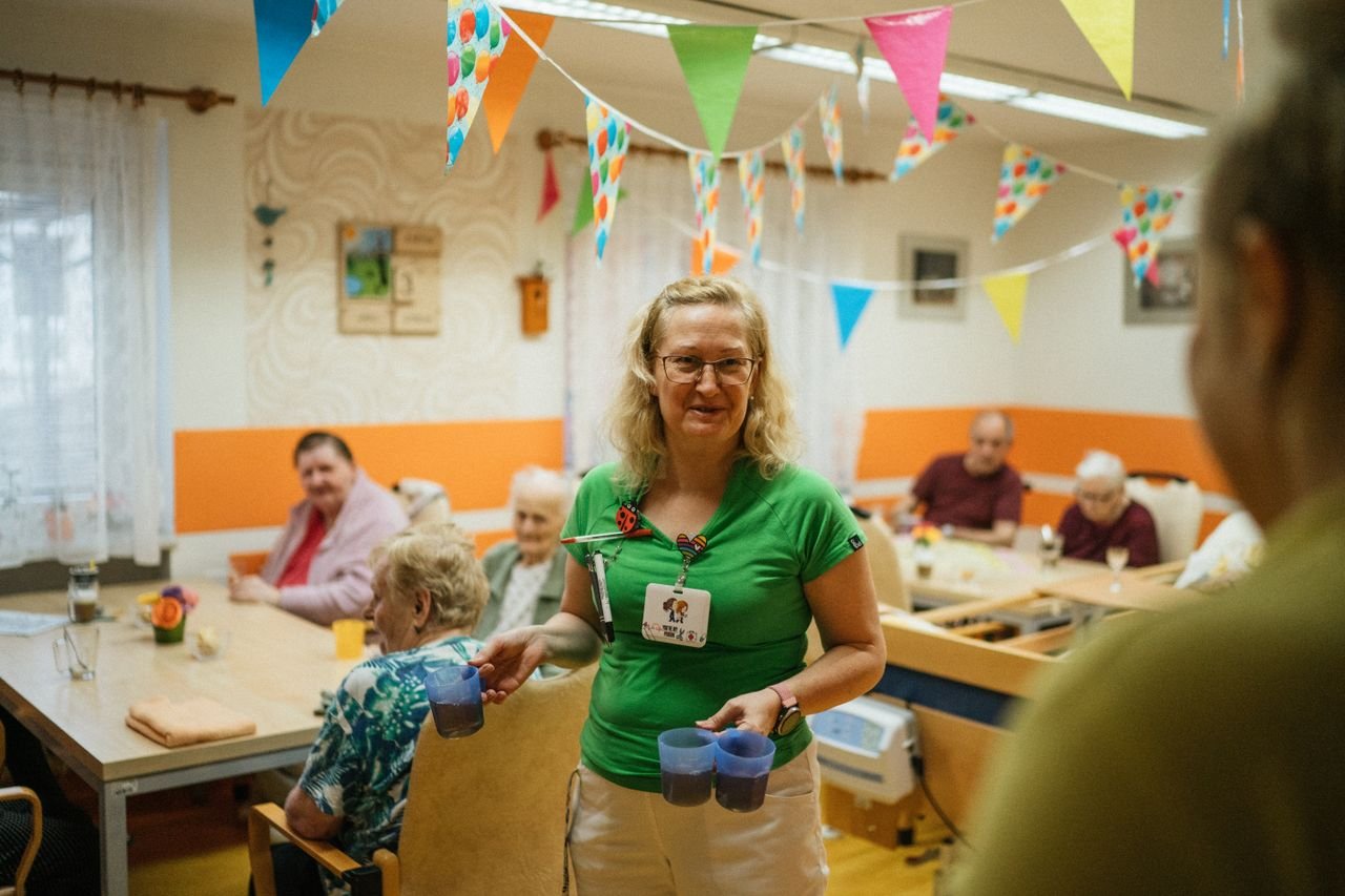 about-04 A caregiver serving drinks to elderly residents in a cheerful nursing home setting.